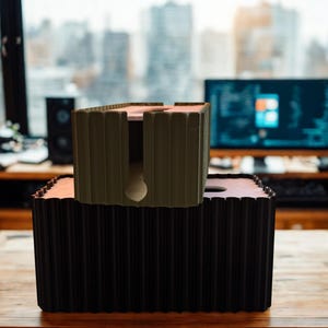 May include: A large black rectangular cable management box with a ribbed exterior texture and a circular cutout on top, topped by a smaller olive green cable management box featuring a similar ribbed design and a central vertical slot.