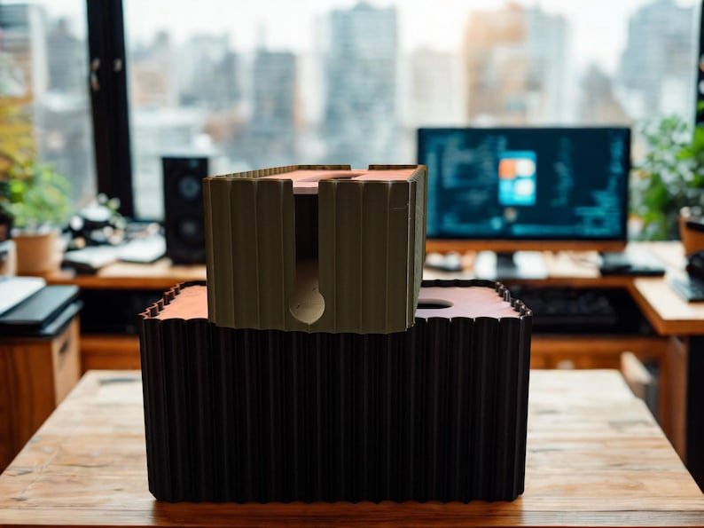 May include: A large black rectangular cable management box with a ribbed exterior texture and a circular cutout on top, topped by a smaller olive green cable management box featuring a similar ribbed design and a central vertical slot.