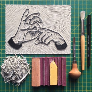 May include: A linocut print of hands, with various printmaking tools. Includes a brush, a carving tool, a black marker, a block of pink and yellow ink, and a pile of cut linoleum. The background is a green grid.