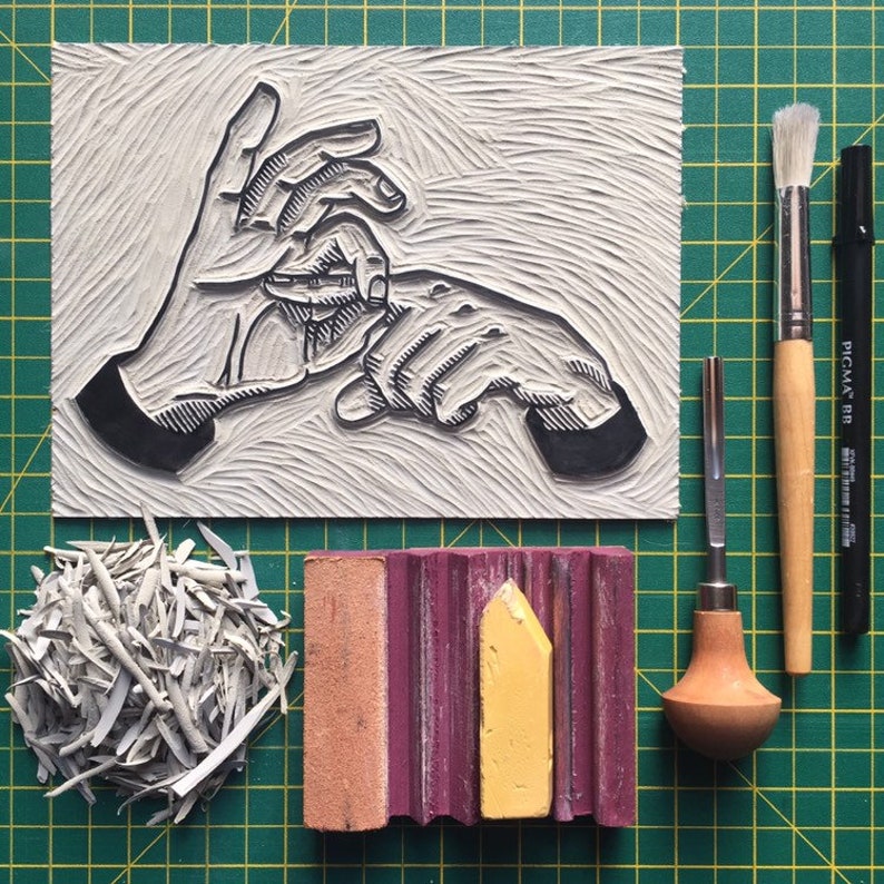 May include: A linocut print of hands, with various printmaking tools. Includes a brush, a carving tool, a black marker, a block of pink and yellow ink, and a pile of cut linoleum. The background is a green grid.