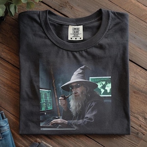Może przedstawiać: Ciemnoszara koszulka z wizerunkiem czarodzieja z długą białą brodą, kapeluszem i okularami przeciwsłonecznymi, palącego fajkę podczas korzystania z komputera. Czarodziej siedzi przed dwoma ekranami komputera. Koszulka ma metkę Comfort Colors.