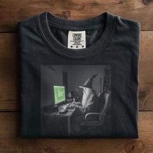 May include: Dark gray t-shirt featuring a graphic of a wizard in a pointed hat, smoking a pipe, and working on a computer. The computer screen displays green text. The shirt has a Comfort Colors tag.