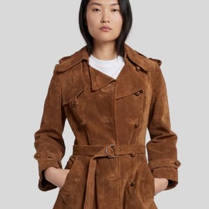 May include: A brown suede trench coat with a double-breasted design, a belt with a circular buckle, and button details. The coat has a classic trench coat collar and pockets. The model is wearing a white t-shirt underneath.