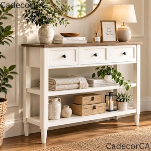 May include: A white wooden console table with a brown top, featuring three drawers with black knobs and two open shelves. The table is styled with a ceramic vase, books, a lamp, a picture frame, woven boxes, a lantern, and potted plants.