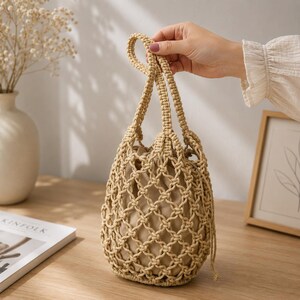 May include: A beige macramé handbag featuring an intricate knotted net design with a solid fabric lining inside. The bag has two matching knotted handles and a drawstring closure, crafted from natural-coloured cord in a structured bucket shape.