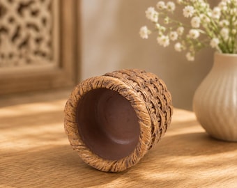 Peut inclure: Un récipient cylindrique doté d'un extérieur texturé imitant un motif de panier tressé dans des tons marron. L'intérieur du récipient est lisse et marron foncé, avec un bord épais en forme de corde entourant l'ouverture supérieure.