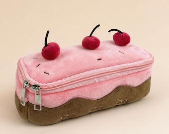 Peut inclure: Une trousse en peluche en forme de gâteau rose avec une base marron. Le gâteau est orné de trois décorations de cerises rouges. La trousse a une fermeture éclair et des tirettes argentées.