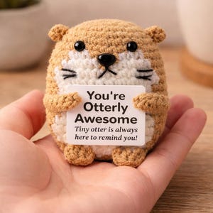 Op de afbeelding: Een klein, rond, gehaakt otter-speeltje gemaakt van beige en wit garen met zwarte plastic ogen en een neus. De otter houdt een wit rechthoekig bordje vast met de zwarte tekst: You're Otterly Awesome. Tiny otter is always here to remind you!