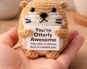 Peut inclure: Un petit jouet loutre crocheté, rond, en fil beige et blanc avec des yeux et un nez en plastique noir. La loutre tient un panneau rectangulaire blanc avec le texte imprimé en noir : You're Otterly Awesome. Tiny otter is always here to remind you!
