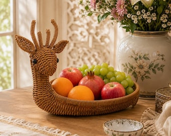 Peut inclure: Un panier à fruits en rotin tressé en forme de cerf avec un long cou et des bois. Le panier contient deux pommes rouges, deux oranges, une grenade et une grappe de raisins verts. La structure en osier brun clair naturel est évidée.