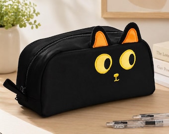 Peut inclure: Trousse noire avec un motif de visage de chat. La trousse présente un corps noir avec des yeux jaunes brodés, un nez et des oreilles orange. Une fermeture éclair noire longe le dessus. La trousse est sur une surface en bois.