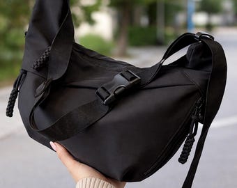 Peut inclure: Sac bandoulière noir avec une bandoulière réglable et une fermeture à boucle. Le sac comporte une poche zippée et des détails décoratifs en cordon tressé. Le sac est tenu dans la main d'une personne.