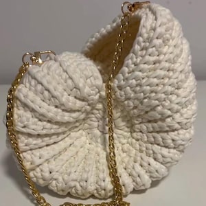 May include: A white, crocheted handbag with a gold chain strap. The bag has a rounded shape with a textured surface. The gold chain is attached to the bag with gold clasps. The bag is sitting on a white surface.