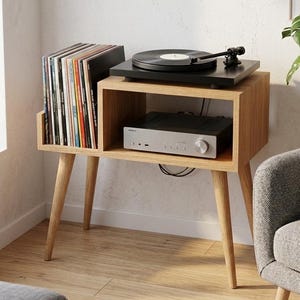 Peut inclure: Un meuble pour platine vinyle en bois avec une étagère intégrée pour un récepteur stéréo et le stockage de disques vinyles. Le meuble a une platine noire sur le dessus et est soutenu par quatre pieds inclinés. Le meuble est dans un salon.