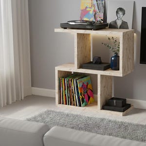 May include: A light-coloured wooden record player stand with a built-in shelf. The stand holds a black record player, vinyl records, and decorative items. The stand is placed in a living room setting with a grey rug and a white couch.