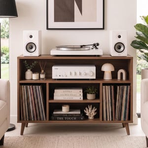 Puede incluir: Un mueble de entretenimiento de estilo moderno con un tocadiscos blanco, altavoces y un amplificador. El mueble de madera marrón tiene estantes llenos de discos de vinilo, libros y objetos decorativos. Una lámpara y dos sofás color crema están en primer plano.