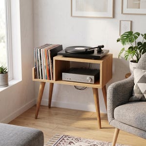 Puede incluir: Un mueble multimedia de madera marrón claro con cuatro patas cónicas, que cuenta con una sección de almacenamiento vertical para discos de vinilo a la izquierda y un estante abierto a la derecha con un amplificador de audio plateado, coronado por un tocadiscos negro.