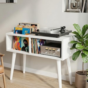 May include: A white wooden media console with four tapered legs, featuring two open storage compartments filled with vinyl records, a black turntable on the top surface, and a silver audio amplifier with three analog gauges inside the right shelf.