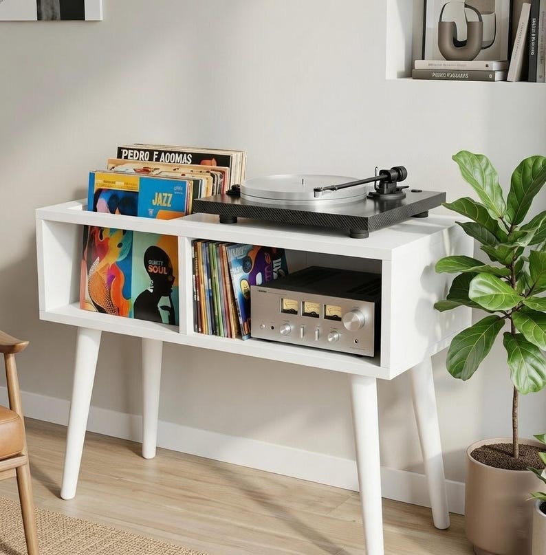 May include: A white wooden media console with four tapered legs, featuring two open storage compartments filled with vinyl records, a black turntable on the top surface, and a silver audio amplifier with three analog gauges inside the right shelf.
