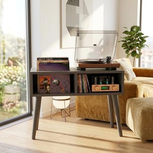 May include: A dark gray wooden record player console with four tapered legs. The top surface holds a wood-grain turntable with a clear dust cover. The lower section features two open storage compartments containing vinyl records and an audio amplifier.