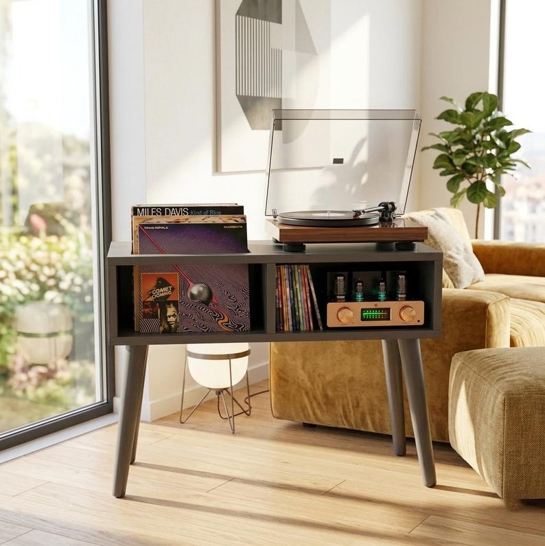 May include: A dark gray wooden record player console with four tapered legs. The top surface holds a wood-grain turntable with a clear dust cover. The lower section features two open storage compartments containing vinyl records and an audio amplifier.