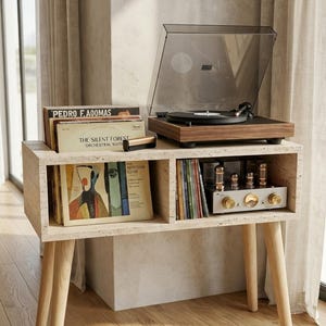 May include: A light-colored stone console table with four wooden legs, featuring a walnut-finish record player on top. The unit has two open shelves holding vinyl records, a cleaning brush, and a silver-toned vacuum tube amplifier with gold knobs.