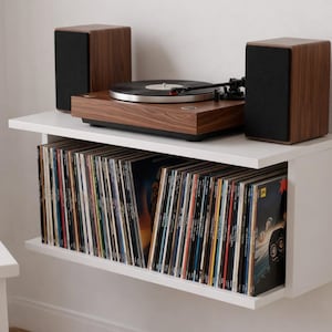 May include: A modern, wall-mounted record player setup. The unit features a wooden turntable with a black record, two wooden speakers, and a lower shelf filled with vinyl records. The unit is white and mounted on a white wall.