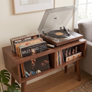 May include: A dark wood record console with tapered legs, featuring a top-mounted turntable with a clear dust cover, a storage compartment filled with vinyl records including Miles Davis, and a lower shelf with an electronic tube amplifier.