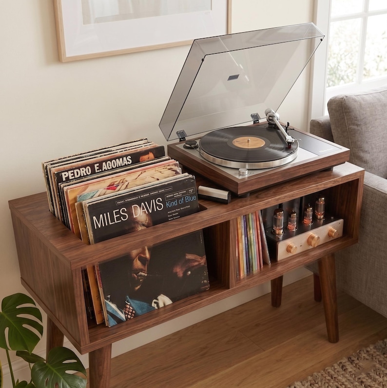 May include: A dark wood record console with tapered legs, featuring a top-mounted turntable with a clear dust cover, a storage compartment filled with vinyl records including Miles Davis, and a lower shelf with an electronic tube amplifier.