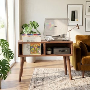 May include: A walnut-toned wooden media console with four tapered legs, featuring an open storage compartment for vinyl records and a shelf holding a silver audio receiver. A clear acrylic turntable sits on top, alongside a small potted succulent.