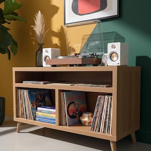 May include: A wooden record player console with a turntable, speakers, and vinyl records. The console is light brown with a mid-century modern design. The speakers are white with gold accents. The records are stored in open compartments.