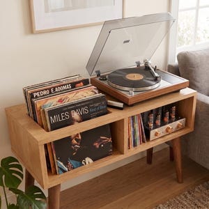 May include: A light wood media console with four tapered legs, featuring a top-mounted turntable with a clear dust cover, a storage compartment filled with vinyl records including Miles Davis Kind of Blue, and a tube amplifier on a lower shelf.