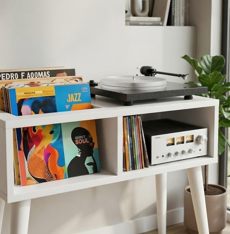 May include: A white wooden media console with four tapered legs, featuring a black turntable on top. The console has open shelving holding vinyl records with covers labeled Jazz and Quiet Soul, and a silver audio amplifier with three gauges.