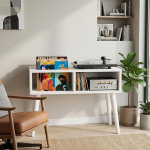 May include: A white rectangular media console with four tapered legs, featuring two open storage compartments. The top surface holds a black turntable. The left shelf contains vinyl records, while the right shelf holds a silver audio receiver.