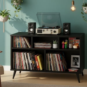 Può includere: Una console nera per dischi in vinile con un giradischi color crema, due altoparlanti neri e ripiani pieni di dischi in vinile. La console è contro un muro turchese, con piante sospese e una lampada.