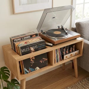 Puede incluir: Consola de tocadiscos de madera con tocadiscos, tapa abierta y una colección de discos de vinilo. La consola tiene dos compartimentos de almacenamiento, uno para discos y otro para equipos de audio. La consola es de madera clara.