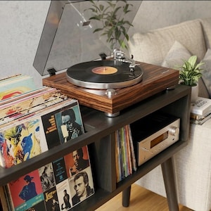 May include: A dark wood grain turntable with a clear dust cover sits on a black console table. A black vinyl record is on the platter. Below, a silver audio receiver and multiple vinyl record sleeves are organized in the storage compartments.