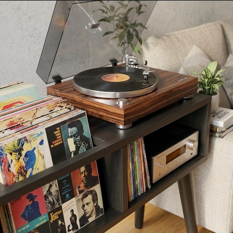 May include: A dark wood grain turntable with a clear dust cover sits on a black console table. A black vinyl record is on the platter. Below, a silver audio receiver and multiple vinyl record sleeves are organized in the storage compartments.