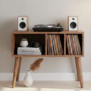 May include: A wooden record player console with a turntable, two speakers, and storage for vinyl records. The console has a mid-century modern design with angled legs. A white vase with dried grass and a small succulent plant are also visible.
