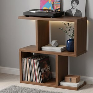 Può includere: Un tavolino in legno marrone con un ripiano integrato e un giradischi sopra. Il tavolo ha un design moderno con luce integrata. Il ripiano contiene dischi in vinile, libri e oggetti decorativi. Il tavolo è alto 76 cm.