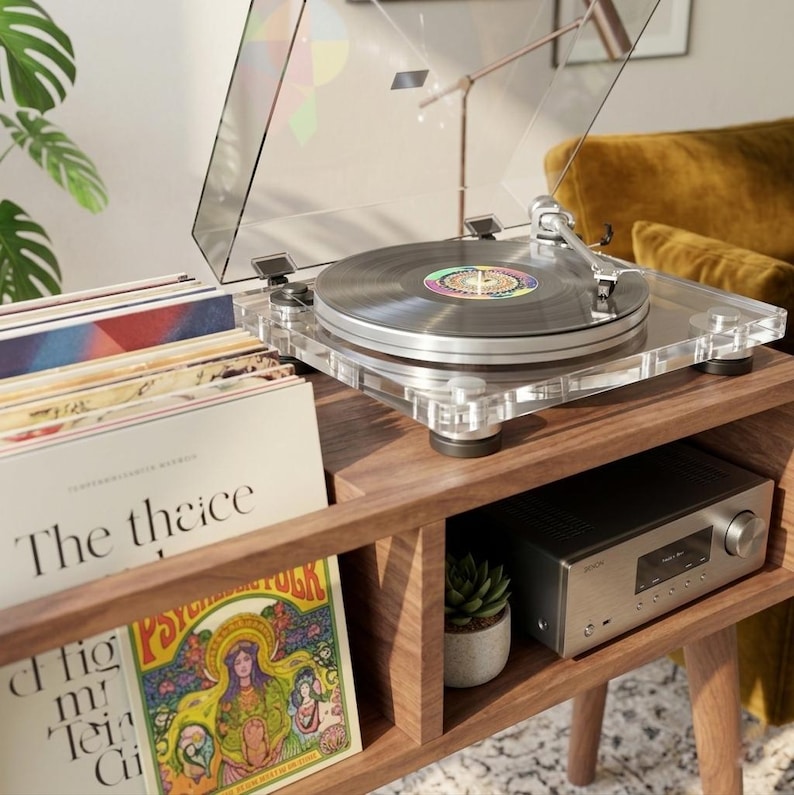 May include: A clear acrylic turntable with a silver platter and tonearm sits on a wooden console. Beside it, a stack of vinyl records includes one with the text The thaice and another with a colorful illustration titled Psychedelic Folk.