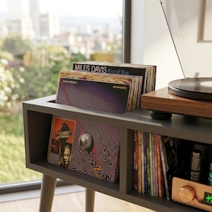 May include: A dark gray wooden vinyl record storage console with a top slot holding several records, including Miles Davis Kind of Blue and The Comet Is Coming. A turntable sits on the right side, and a shelf below holds additional records.