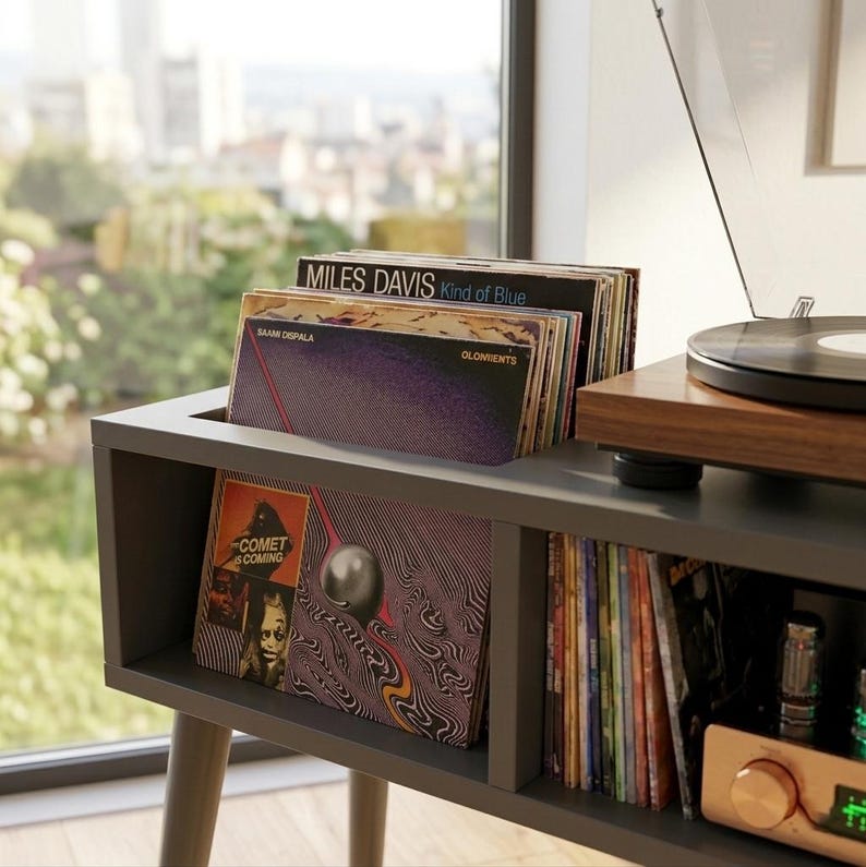 May include: A dark gray wooden vinyl record storage console with a top slot holding several records, including Miles Davis Kind of Blue and The Comet Is Coming. A turntable sits on the right side, and a shelf below holds additional records.
