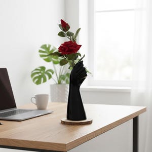 May include: A black ceramic vase shaped like a hand holding a red rose and bud. The vase sits on a wooden coaster on a light-colored wooden desk. A laptop and a white mug are also on the desk.