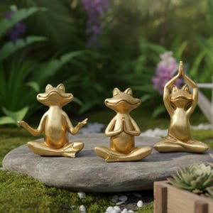 May include: Three gold-colored frog figurines in yoga poses. The frogs are seated on a gray stone, with a lush green and purple background. One frog has arms outstretched, another has hands together, and the third has arms raised.