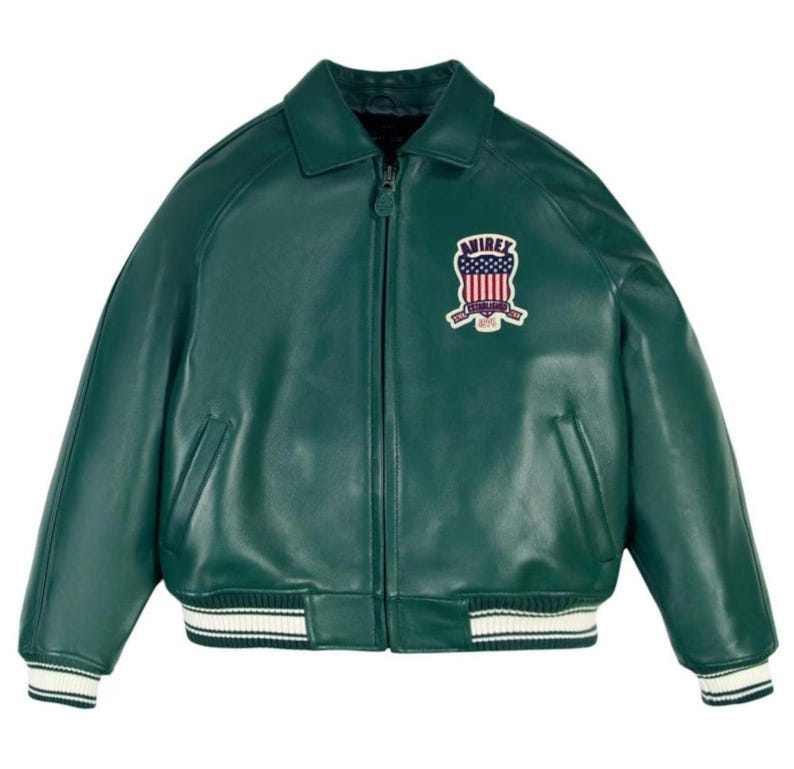 May include: A dark green leather jacket with a zip-up front, collar, and a patch on the chest. The patch features a design with the text "AVIREX" and an American flag. The jacket has white and black striped cuffs and hem.