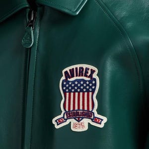 May include: Close-up of a dark green leather jacket with a zipper and a collar. The jacket features a patch with the word "AVIREX" above a design of the American flag and the words "ESTABLISHED NY 1975".
