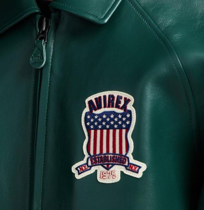 May include: Close-up of a dark green leather jacket with a zipper and a collar. The jacket features a patch with the word "AVIREX" above a design of the American flag and the words "ESTABLISHED NY 1975".