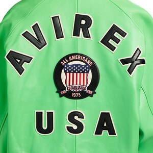 May include: Close-up of a bright green jacket with black and white lettering and patches. The jacket features the word "AVIREX" across the top and "USA" at the bottom. A circular patch in the center reads "ALL AMERICANS ESTABLISHED 1975".