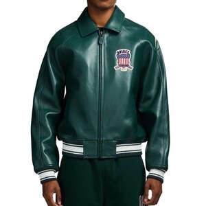 May include: A dark green leather jacket with a zip-up front and a collar. The jacket features a white and black striped band at the waist and cuffs. An embroidered patch with the text "Avirex" and a patriotic design is on the chest.